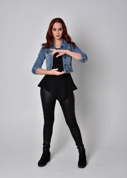  portrait of a pretty girl with red hair wearing black leather pants and denim jacket. Full length standing pose isolated against a  grey studio background - Photo, Image