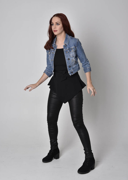  portrét hezké dívky s rudými vlasy v černých kožených kalhotách a džínové bundě. Plná délka stojící póza izolované na šedém pozadí studia - Fotografie, Obrázek