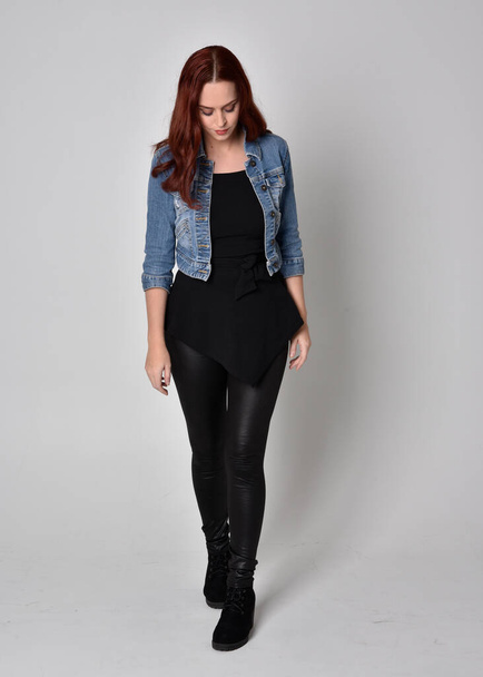  portrait of a pretty girl with red hair wearing black leather pants and denim jacket. Full length standing pose isolated against a  grey studio background - Valokuva, kuva