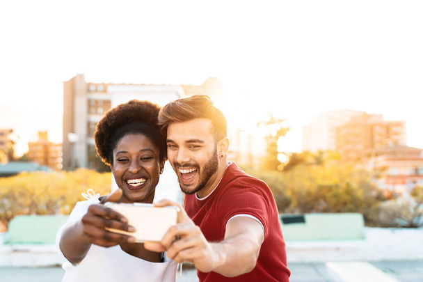 Happy couple taking selfie with mobile smartphone outdoor - Mixed race friends having fun with new trendy technology outdoor - Youth millennial generation and multi ethnic teenagers lifestyle concept - Photo, Image