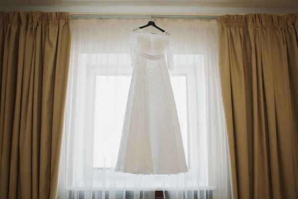 Νυφικό φόρεμα της νύφης ζυγίζει σε μια ξύλινη κρεμάστρα. Κρεμ φόρεμα. Το πρωινό της νύφης. Τελετή επίσκεψης. Νύφη και γαμπρός. Γάμος. ? φόρεμα ocktail, βραδινό φόρεμα. - Φωτογραφία, εικόνα