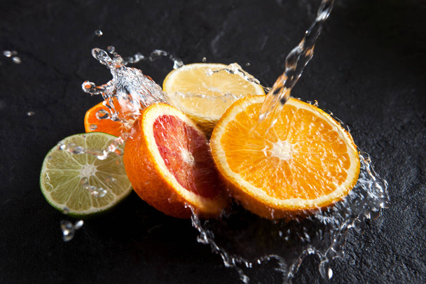 Water pours into halves of fresh ripe citrus fruits on a dark stone background. Splashes fall on red orange, lemon and lime. Selective focus. Close-up, top view. - Фото, зображення