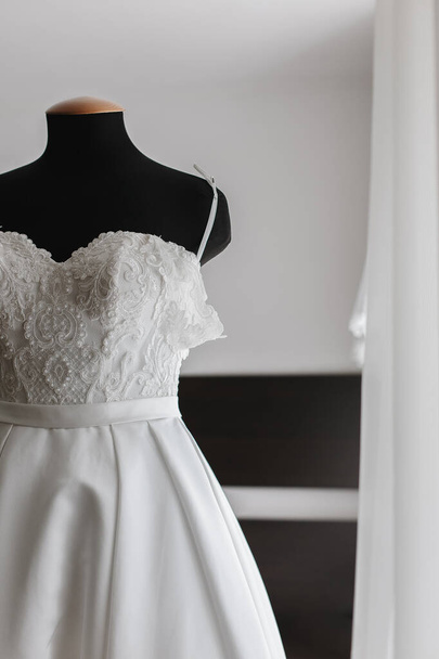Νυφικό φόρεμα της νύφης ζυγίζει σε μια ξύλινη κρεμάστρα. Κρεμ φόρεμα. Το πρωινό της νύφης. Τελετή επίσκεψης. Νύφη και γαμπρός. Γάμος. ? φόρεμα ocktail, βραδινό φόρεμα. - Φωτογραφία, εικόνα