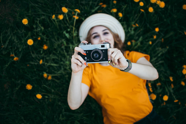 Junges schönes Mädchen in orangefarbenem T-Shirt mit Retro-Kamera in der Hand auf dem Rasen liegend, wo Löwenzahn wächst, blühender Löwenzahn. - Foto, Bild