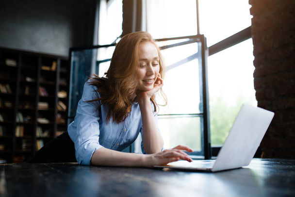 Redhead businesswoman put her chin on her hand and looks pretty at the laptop screen while doing her job. - Foto, imagen