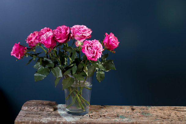 Bouquet of red roses in a glass vase on a dark blue background on a wooden wrecked surface of the bench. Copy space - Φωτογραφία, εικόνα