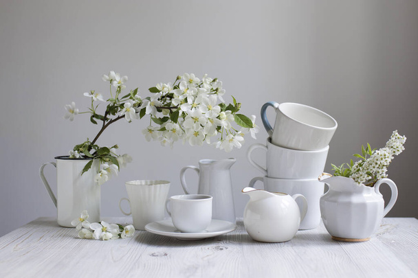 Vase with a blossoming branch of an apple tree. A set of white dishes: a cup of coffee, milk jug, a jug, a tray on the table against the background of a white wall. Copy space - Foto, Bild