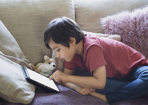 Çocuk ödevi için tablet kullanıyor, çocuk kanepede uzanıyor çizgi film seyrediyor ya da dijital tablette oyun oynuyor, evde eğitim, sosyal uzaklık, e-öğrenim online eğitim - Fotoğraf, Görsel