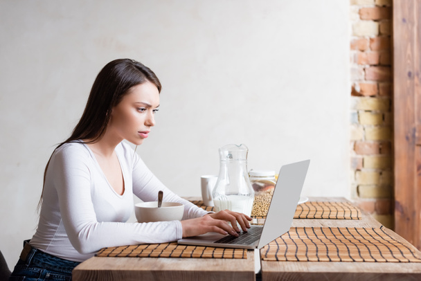 όμορφη freelancer χρησιμοποιώντας φορητό υπολογιστή κοντά κανάτα με γάλα και μπολ  - Φωτογραφία, εικόνα
