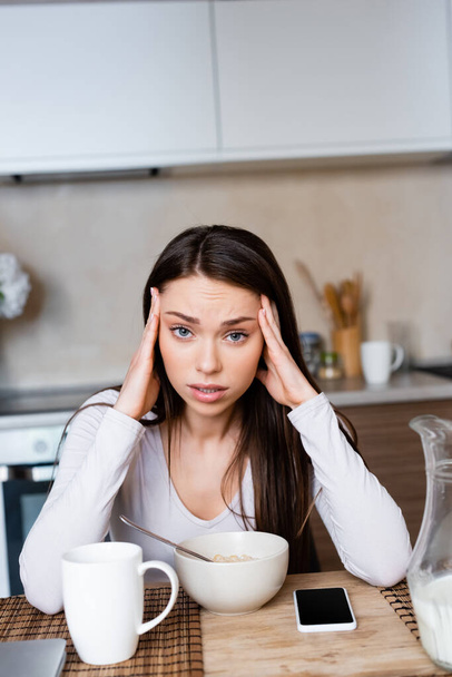 upset girl touching head near bowl, jug and smartphone with blank screen - Foto, Imagen