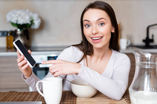 enfoque selectivo de la chica feliz apuntando con el dedo al teléfono inteligente con pantalla en blanco cerca de la taza, tazón y jarra con leche
  - Foto, Imagen