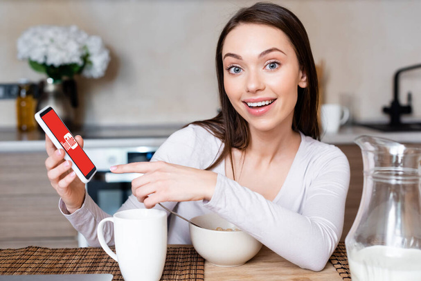 KYIV, UKRAINE - APRIL 29, 2020: selective focus of happy girl pointing with finger at smartphone with youtube app near cup, bowl and jug with milk  - 写真・画像