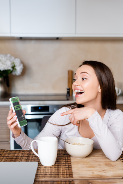 excited girl pointing with finger at smartphone with booking app near cup and bowl  - Foto, imagen