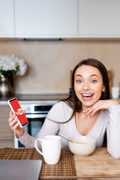 KYIV, UKRAINE - APRIL 29, 2020: excited woman holding smartphone with youtube app near cup and bowl  - 写真・画像