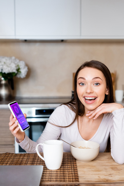 excited woman holding smartphone with online shopping app near cup and bowl  - Фото, изображение