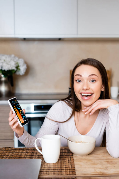 excited woman holding smartphone with charts and graphs near cup and bowl  - 写真・画像