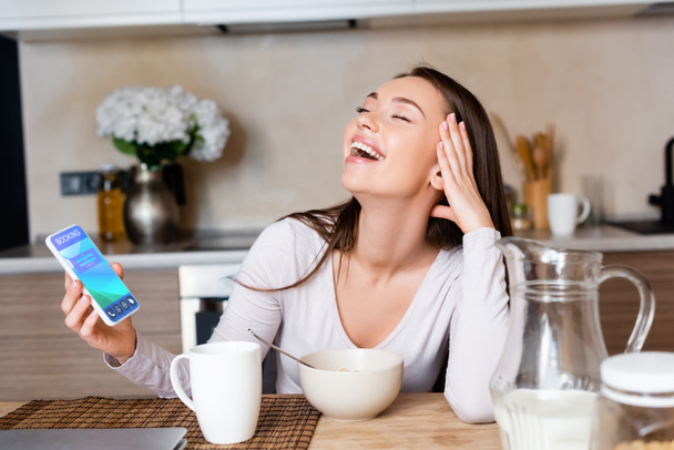 mujer alegre sosteniendo teléfono inteligente con aplicación de reserva y riendo cerca del desayuno
 - Foto, Imagen