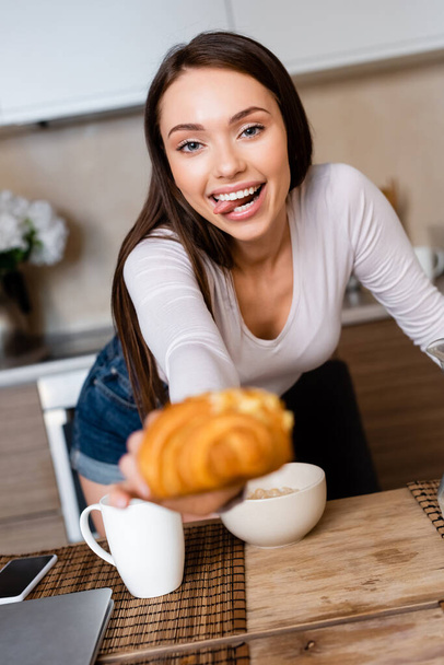 selective focus of girl holding tasty croissant and sticking out tongue - Valokuva, kuva