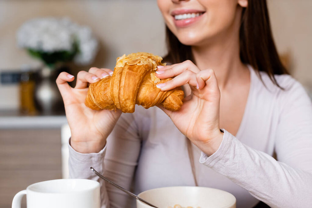 oříznutý pohled na šťastnou dívku drží chutný croissant v blízkosti misky a poháru  - Fotografie, Obrázek