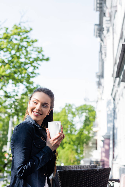 Χαμογελαστή νεαρή γυναίκα με τζιν μπουφάν που κρατάει καφέ απ 'έξω.  - Φωτογραφία, εικόνα