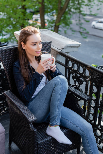 glückliches Mädchen in Jeans und Jacke, das auf einem Stuhl sitzt und eine Tasse Kaffee hält  - Foto, Bild
