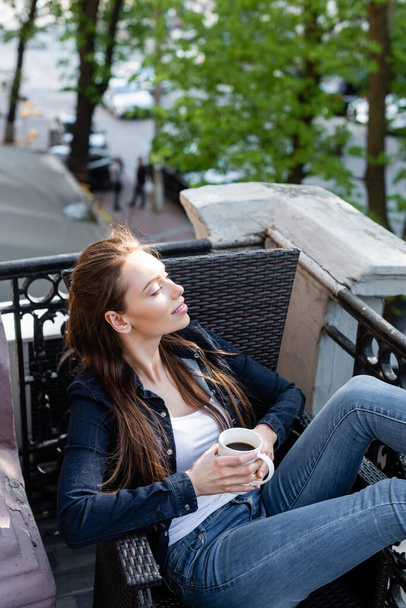 cheerful girl in denim jeans and jacket sitting on chair and holding cup of coffee on balcony  - Fotó, kép
