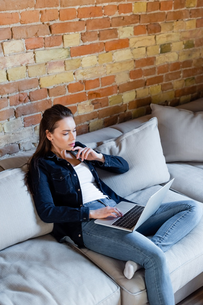 pensive freelancer using laptop while sitting on sofa  - Photo, Image