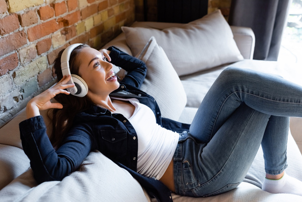 cheerful girl touching wireless headphones while listening music  - Фото, изображение
