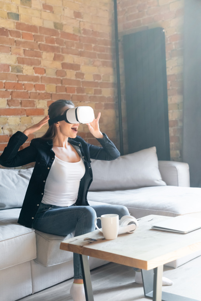 izgatott fiatal nő megérinti virtuális valóság headset, miközben ül a kanapén - Fotó, kép