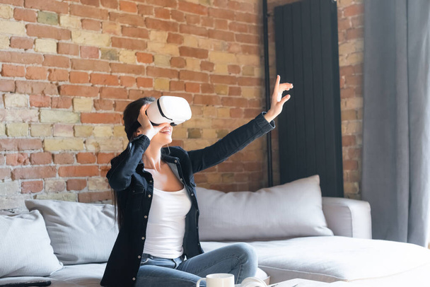 happy young woman touching virtual reality headset and gesturing while sitting on sofa - Photo, Image