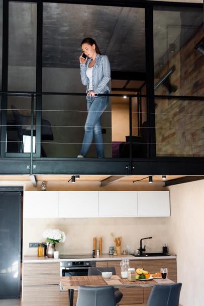 young woman talking on smartphone while standing on second floor in duplex apartment  - Fotografie, Obrázek