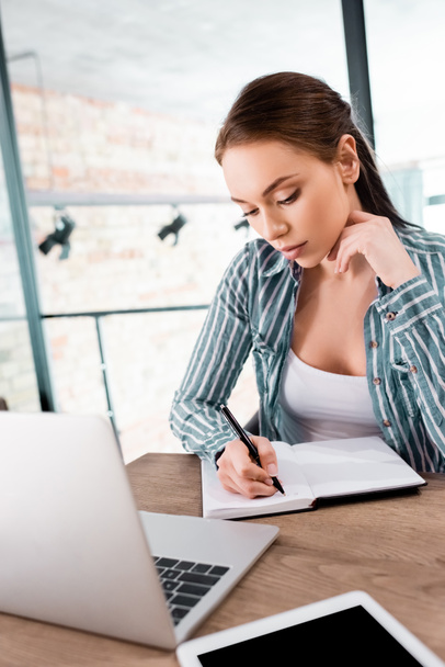 selective focus of beautiful woman writing in notebook near laptop and digital tablet with blank screen, online study concept - Фото, изображение