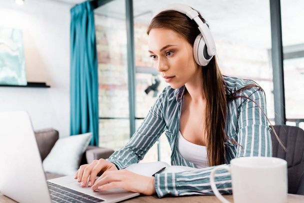 selective focus of beautiful freelancer in wireless headphones using laptop near cup - Foto, Imagen