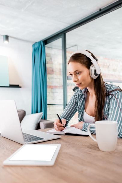 selektywne skupienie dziewczyny w słuchawkach bezprzewodowych pisanie w notebooku w pobliżu laptopa i tabletu cyfrowego z białym ekranem, koncepcja badania online - Zdjęcie, obraz