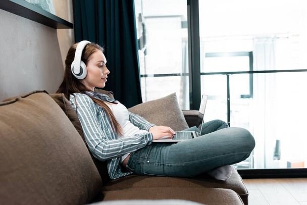 side view of attractive freelancer in wireless headphones using laptop in living room  - Фото, изображение