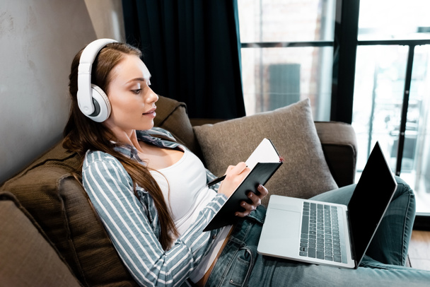attractive woman in wireless headphones writing in notebook near laptop with blank screen, online study concept  - Foto, imagen