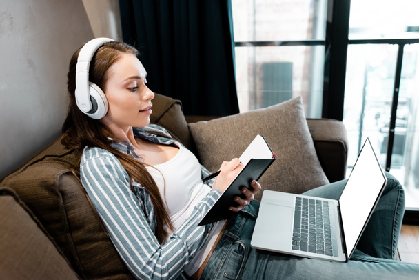 attraktive Frau mit drahtlosem Kopfhörer, die in einem Notizbuch in der Nähe von Laptop mit weißem Bildschirm schreibt, Online-Studienkonzept  - Foto, Bild
