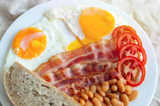Завершує традиційний англійський сніданок: смажені яйця, бекон, квасоля, хліб і помідор на тарілці.. - Фото, зображення