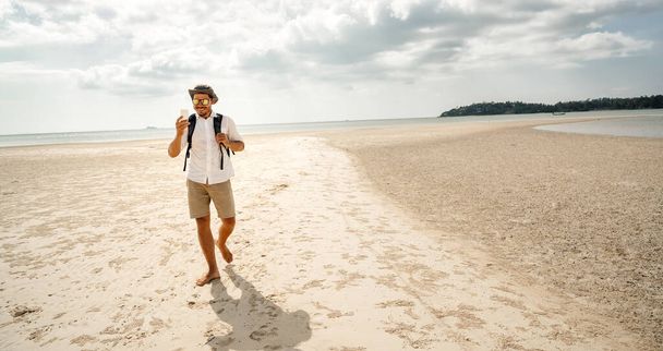 Şapkalı ve gözlüklü genç adam tropikal bir adanın ıssız bir sahilinde elinde akıllı bir telefonla selfie karesi yapıyor. Seyahat ve mobil iletişim. - Fotoğraf, Görsel