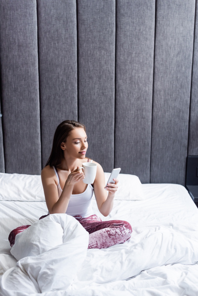 bela jovem mulher usando smartphone e segurando xícara de café na cama
  - Foto, Imagem