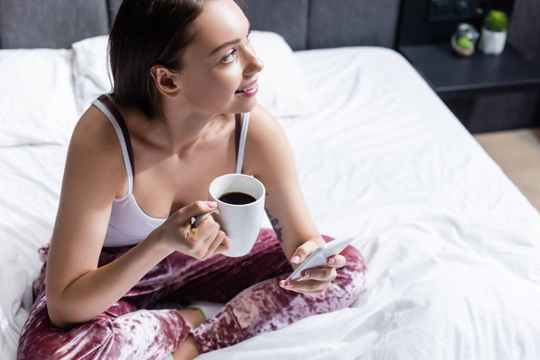 happy young woman holding smartphone and cup of coffee in bed  - 写真・画像