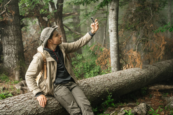 άνδρας με ρούχα πεζοπορίας κρατά ένα smartphone στο χέρι του, ενώ κάθεται σε ένα κούτσουρο στο δάσος και βγάζει μια φωτογραφία selfie ή χρησιμοποιεί κινητή επικοινωνία για να συνομιλήσει στο διαδίκτυο. Άνθρωπος και πλοηγός. - Φωτογραφία, εικόνα