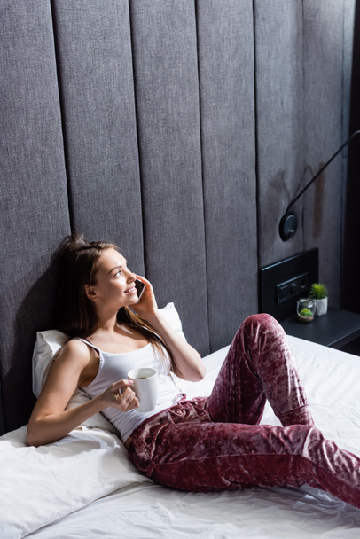 happy woman talking on smartphone and holding cup of coffee in bed - Photo, Image