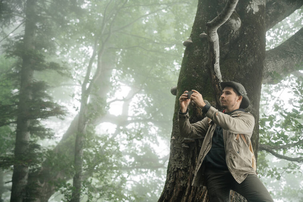 Человек в костюме и шляпе делает селфи на смартфоне возле дерева в туманном лесу. Одинокие прогулки и прогулки по лесу
. - Фото, изображение