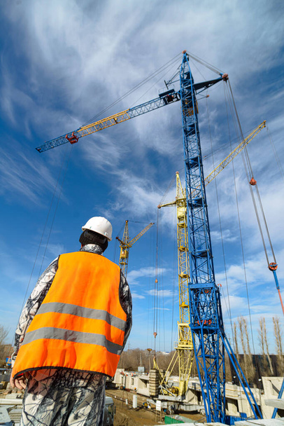 Строительство дома. Рабочий в комбинезоне смотрит на высотные строительные краны на строительной площадке против голубого неба. Фотография снизу вверх на широкоугольном объективе
 - Фото, изображение