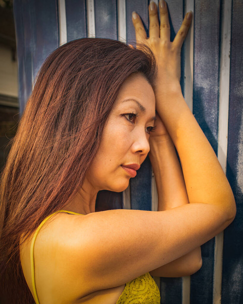 Pensive traurig aussehende reife asiatische Frau mit langen Haaren auf einem gestreiften dunkelblauen Hintergrund. - Foto, Bild