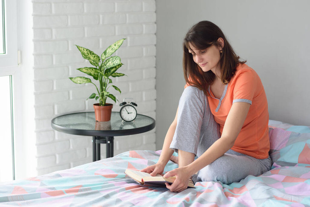 Κορίτσι που κάθεται στο κρεβάτι με πιτζάμες και διαβάζει ένα βιβλίο - Φωτογραφία, εικόνα