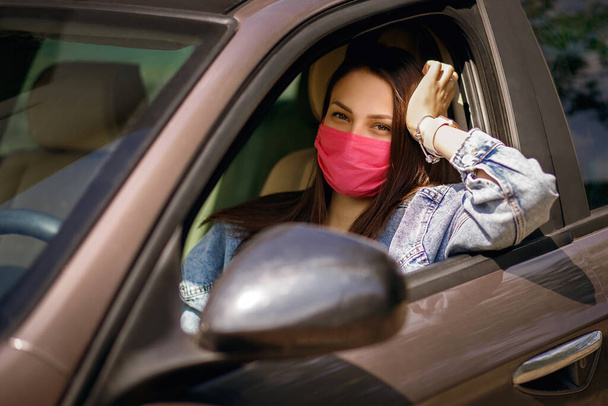 Gyönyörű fiatal lány rózsaszín maszkban ül egy autóban, egy védőmaszk a koronavírustól, egy sofőr a város utcáján a koronavírus kitörésekor, kovid-19, naplemente. - Fotó, kép