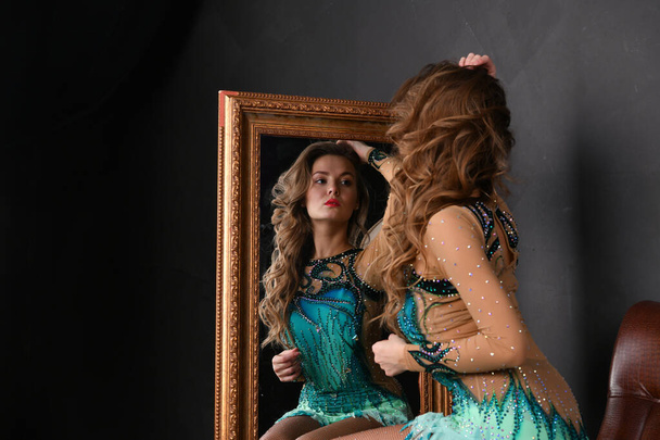 gymnast in a green suit with a golden ball sits in a brown chair and looks in the mirror - Photo, Image
