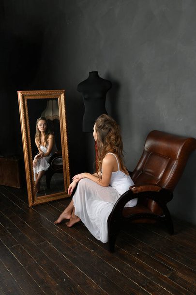 ballerina in een witte jurk en rode punten zit in een bruine stoel en kijkt in de spiegel - Foto, afbeelding